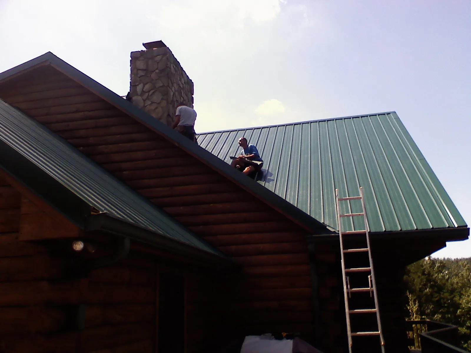 Roofers installing green metal panels beside a stone chimney on a log home in Lancaster